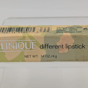 May include: A Clinique lipstick box with a camouflage pattern in shades of green, tan, and beige. The box features the Clinique logo, the words "different lipstick," and the net weight of .14 oz./4g.