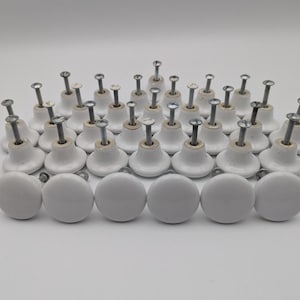 May include: A collection of thirty-two white ceramic cabinet knobs, each featuring a smooth, rounded top and a silver-toned metal screw extending from the back. The knobs are arranged in a grid-like formation, showcasing their uniform design.