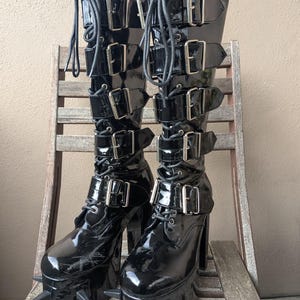 May include: Pair of black patent leather platform boots with multiple buckles and laces. The boots have a chunky heel and a platform sole with metal spikes. The boots are sitting on a wooden chair.