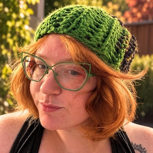 May include: A hand-knitted beanie featuring a vibrant green yarn with a textured, ribbed pattern. The hat has a slouchy fit and transitions into a darker, black-toned yarn at the back. The knit construction is thick, showing clear loops.