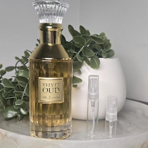 May include: A gold-coloured perfume bottle with a clear, faceted stopper and the words "Velvet Oud" on the label. Two small, clear spray bottles are next to the perfume bottle. The perfume bottle and spray bottles are on a marble tray.