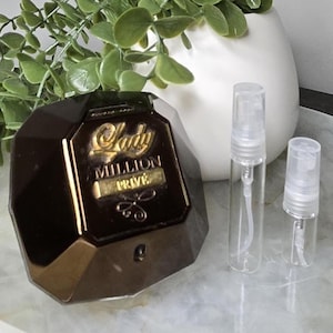 May include: A dark brown perfume bottle with gold lettering reading "Lady Million Privé" sits on a marble tray alongside two small, clear spray bottles. A white vase with green leafy plant stems is in the background.