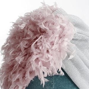 Puede incluir: Primer plano de un turbante con una base de tela plateada brillante y un acento de flecos rosa claro y esponjoso. El turbante est&aacute; sobre un fondo turquesa, creando un contraste de colores y texturas. El dise&ntilde;o es elegante y llamativo.