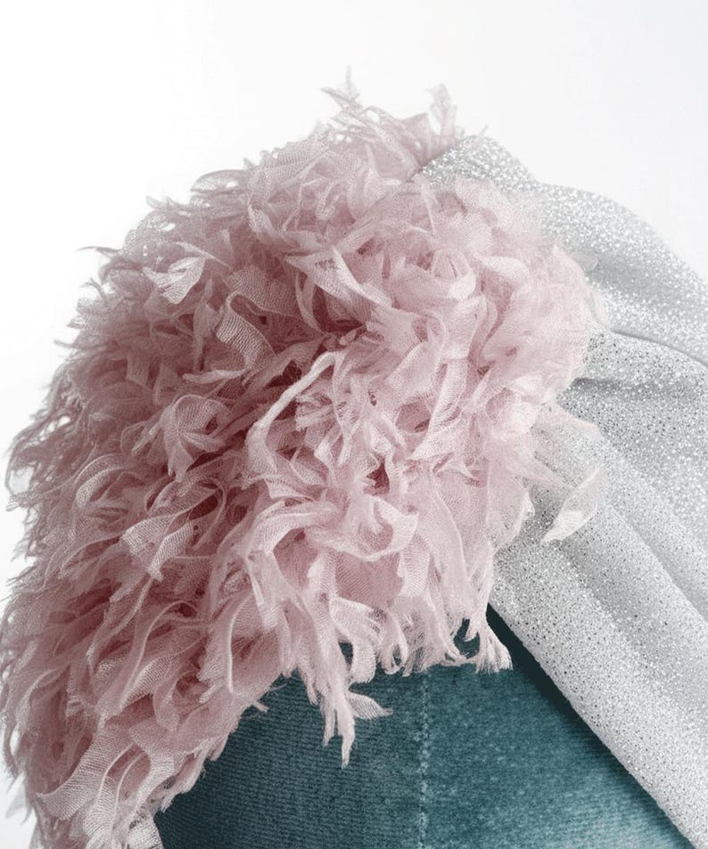 Puede incluir: Primer plano de un turbante con una base de tela plateada brillante y un acento de flecos rosa claro y esponjoso. El turbante est&aacute; sobre un fondo turquesa, creando un contraste de colores y texturas. El dise&ntilde;o es elegante y llamativo.