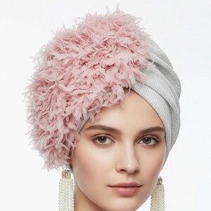 Puede incluir: Un turbante plateado con un adorno rosa y plumoso. La persona lleva pendientes largos de perlas. Este tocado es un accesorio de moda, ideal para ocasiones especiales o para realzar un atuendo.