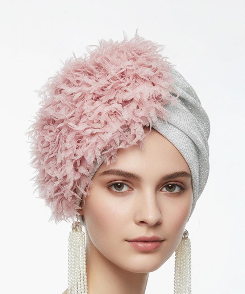Puede incluir: Un turbante plateado con un adorno rosa y plumoso. La persona lleva pendientes largos de perlas. Este tocado es un accesorio de moda, ideal para ocasiones especiales o para realzar un atuendo.