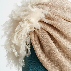 Puede incluir: Un turbante dorado con un dise&ntilde;o de tela texturizada. El turbante est&aacute; adornado con un grupo de plumas blancas y un collar de perlas. El tocado es probablemente para ocasiones especiales.