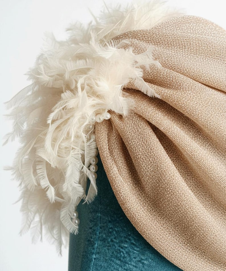 Puede incluir: Un turbante dorado con un dise&ntilde;o de tela texturizada. El turbante est&aacute; adornado con un grupo de plumas blancas y un collar de perlas. El tocado es probablemente para ocasiones especiales.