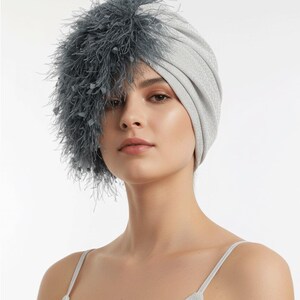 Puede incluir: Un turbante plateado con un adorno de plumas. El tocado est&aacute; hecho de una tela brillante y texturizada. El adorno de plumas es de color gris oscuro y cubre el lado izquierdo del tocado.