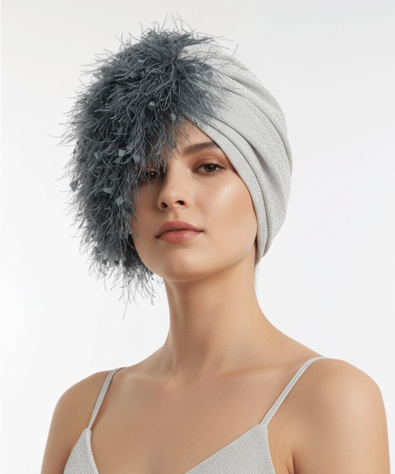 Puede incluir: Un turbante plateado con un adorno de plumas. El tocado est&aacute; hecho de una tela brillante y texturizada. El adorno de plumas es de color gris oscuro y cubre el lado izquierdo del tocado.