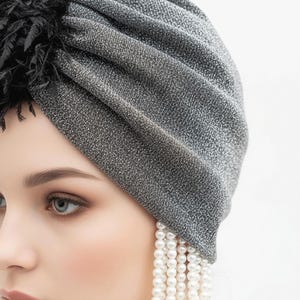 Puede incluir: Un turbante gris plateado con una tela texturizada y brillante. El turbante tiene un detalle de plumas negras y se combina con un pendiente largo de perlas colgantes. Un accesorio para la cabeza elegante.