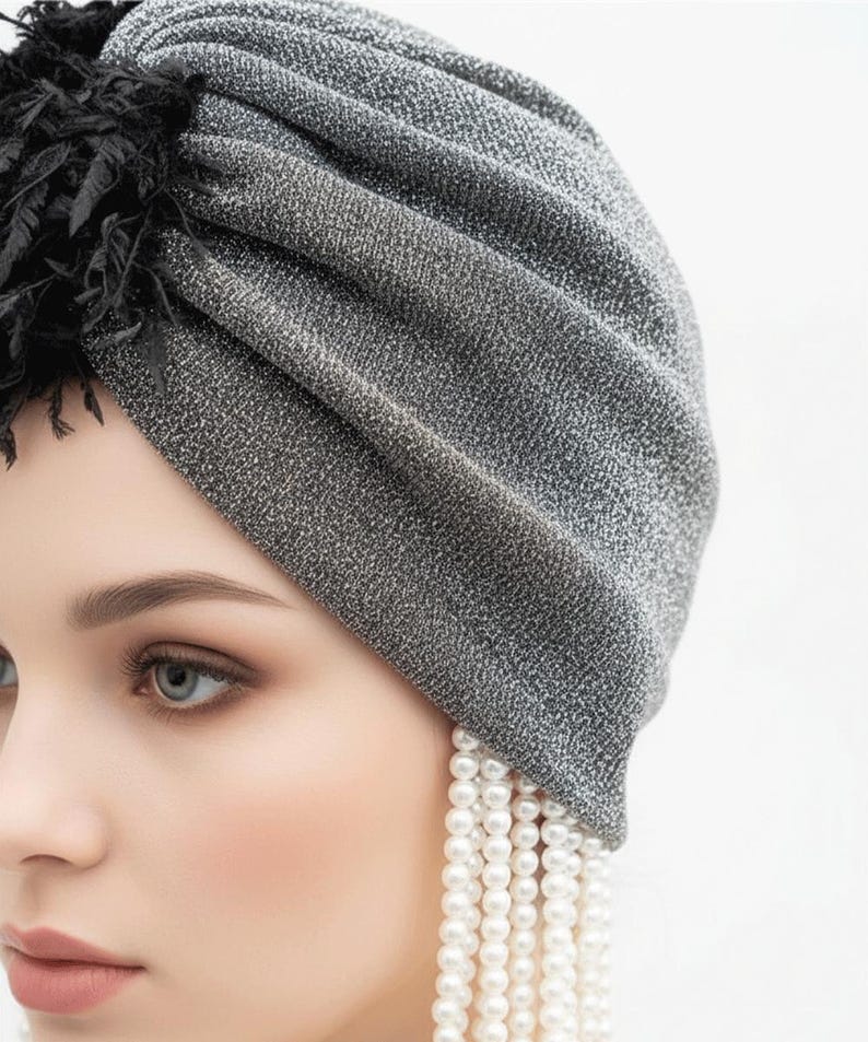 Puede incluir: Un turbante gris plateado con una tela texturizada y brillante. El turbante tiene un detalle de plumas negras y se combina con un pendiente largo de perlas colgantes. Un accesorio para la cabeza elegante.