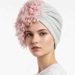 Puede incluir: Un turbante plateado con un adorno de plumas rosas. El turbante est&aacute; hecho de una tela brillante y tiene un dise&ntilde;o fruncido. Las plumas rosas est&aacute;n agrupadas en un lado, agregando un toque de elegancia al tocado.