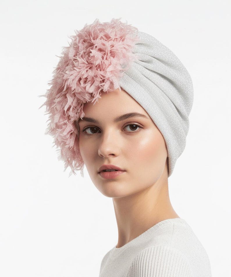 Puede incluir: Un turbante plateado con un adorno de plumas rosas. El turbante est&aacute; hecho de una tela brillante y tiene un dise&ntilde;o fruncido. Las plumas rosas est&aacute;n agrupadas en un lado, agregando un toque de elegancia al tocado.