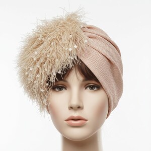 Puede incluir: Un turbante rosa p&aacute;lido con una tela texturizada. El sombrero presenta un gran detalle de plumas en el lado izquierdo, adornado con peque&ntilde;os adornos blancos en forma de cuadrado. Este tocado est&aacute; dise&ntilde;ado para la moda y eventos especiales.