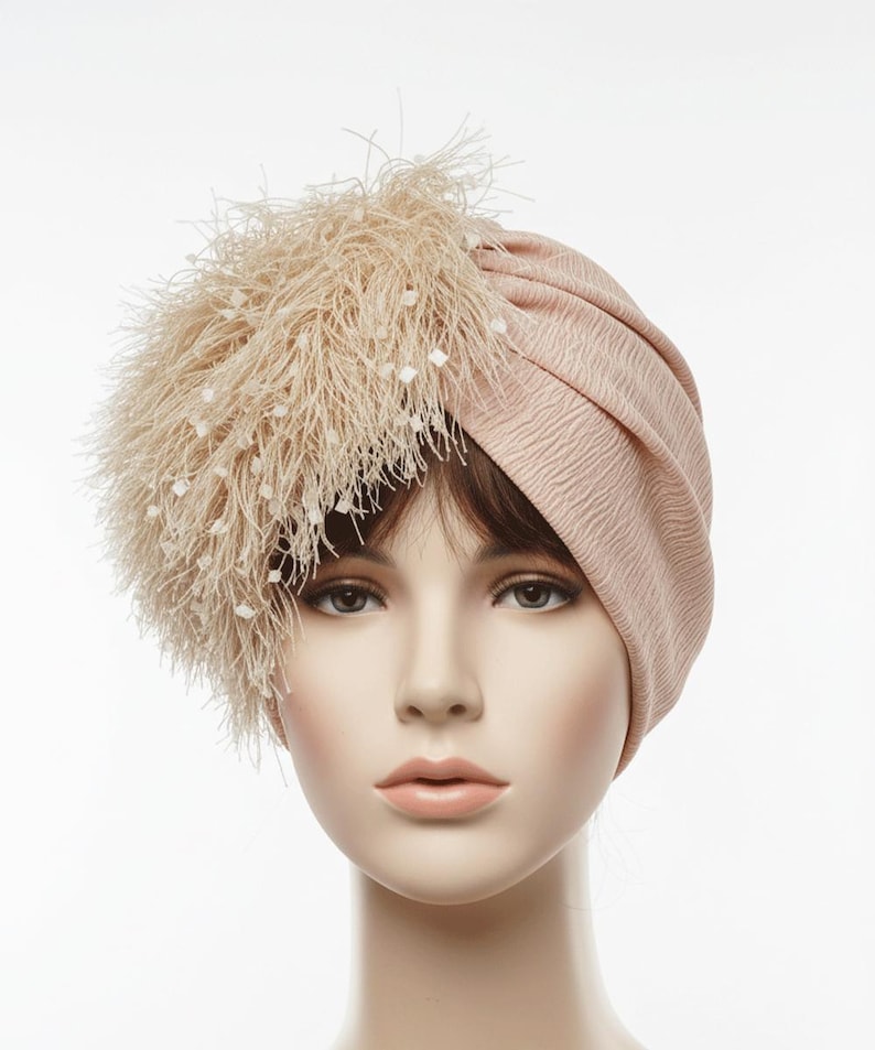 Puede incluir: Un turbante rosa p&aacute;lido con una tela texturizada. El sombrero presenta un gran detalle de plumas en el lado izquierdo, adornado con peque&ntilde;os adornos blancos en forma de cuadrado. Este tocado est&aacute; dise&ntilde;ado para la moda y eventos especiales.
