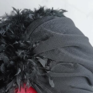 Puede incluir: Un turbante negro de tela texturizada. El turbante est&aacute; adornado con un ribete de plumas negras en el borde superior. El tocado est&aacute; sobre una superficie roja.