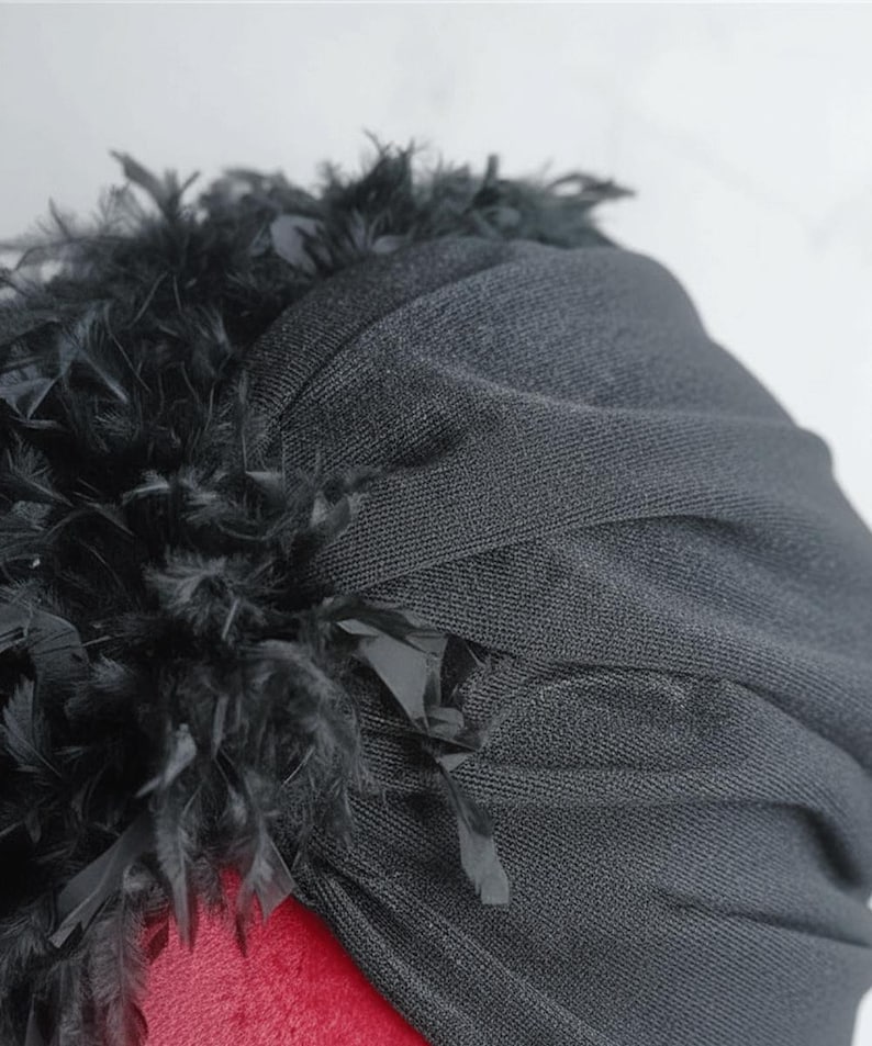 Puede incluir: Un turbante negro de tela texturizada. El turbante est&aacute; adornado con un ribete de plumas negras en el borde superior. El tocado est&aacute; sobre una superficie roja.