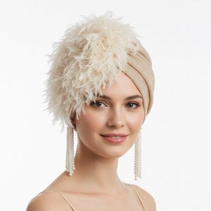 Puede incluir: Un turbante beige con un acento de plumas esponjosas en el lado izquierdo. La mujer lleva pendientes largos de perlas colgantes y una camiseta sin mangas beige a juego. El fondo es blanco.