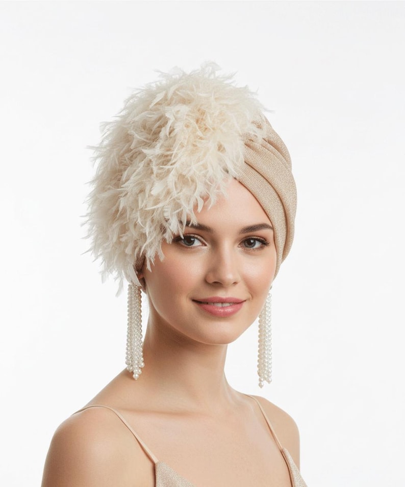 Puede incluir: Un turbante beige con un acento de plumas esponjosas en el lado izquierdo. La mujer lleva pendientes largos de perlas colgantes y una camiseta sin mangas beige a juego. El fondo es blanco.