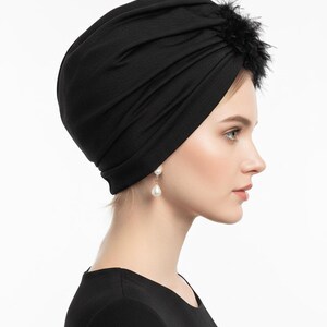 Puede incluir: Un turbante negro con un dise&ntilde;o fruncido. Un grupo de plumas negras adorna la parte superior. El tocado se usa con pendientes de perlas colgantes. El fondo es blanco.