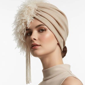 May include: A beige turban-style head covering with a textured appearance. The turban is adorned with a cluster of white feathers on the left side and a strand of pearls. The head covering is worn by a person with a neutral-toned top.