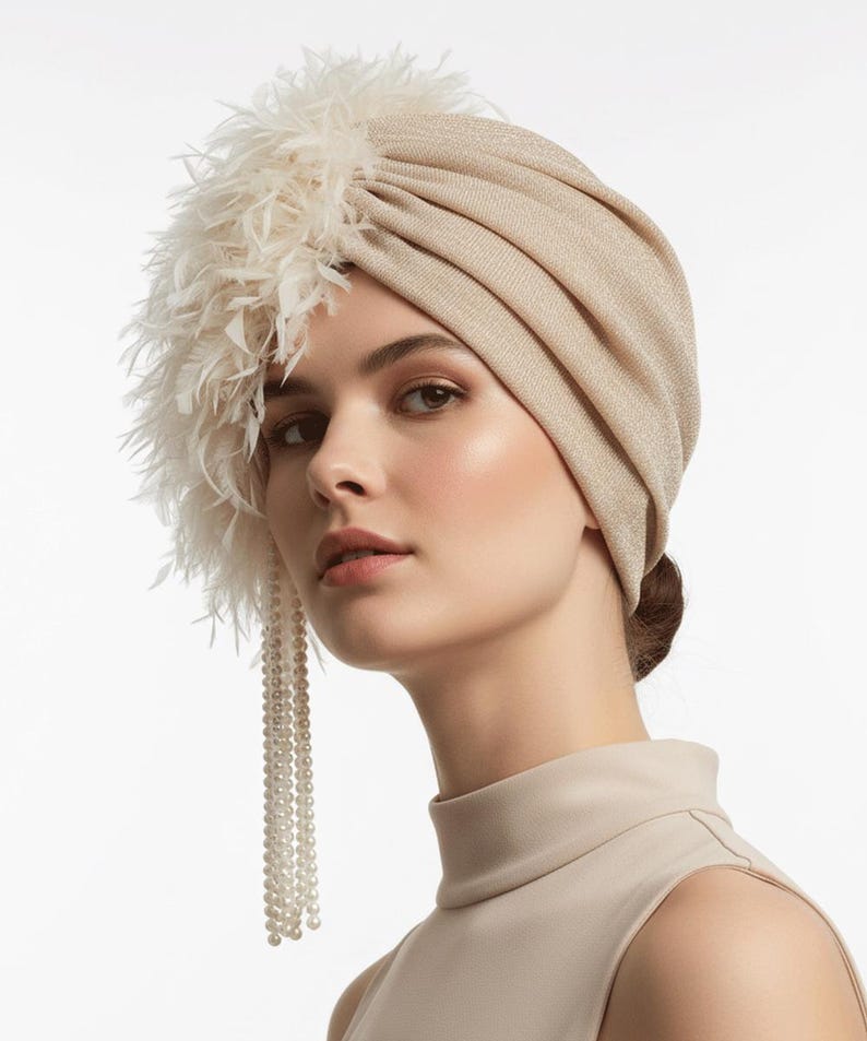Puede incluir: Un turbante beige con aspecto texturizado. El turbante est&aacute; adornado con un grupo de plumas blancas en el lado izquierdo y una hebra de perlas. El tocado lo lleva una persona con un top de tono neutro.