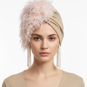 Puede incluir: Un turbante beige con un acento de plumas rosas. La mujer de la imagen lleva pendientes largos de perlas y una blusa beige. El turbante es un tocado elegante.