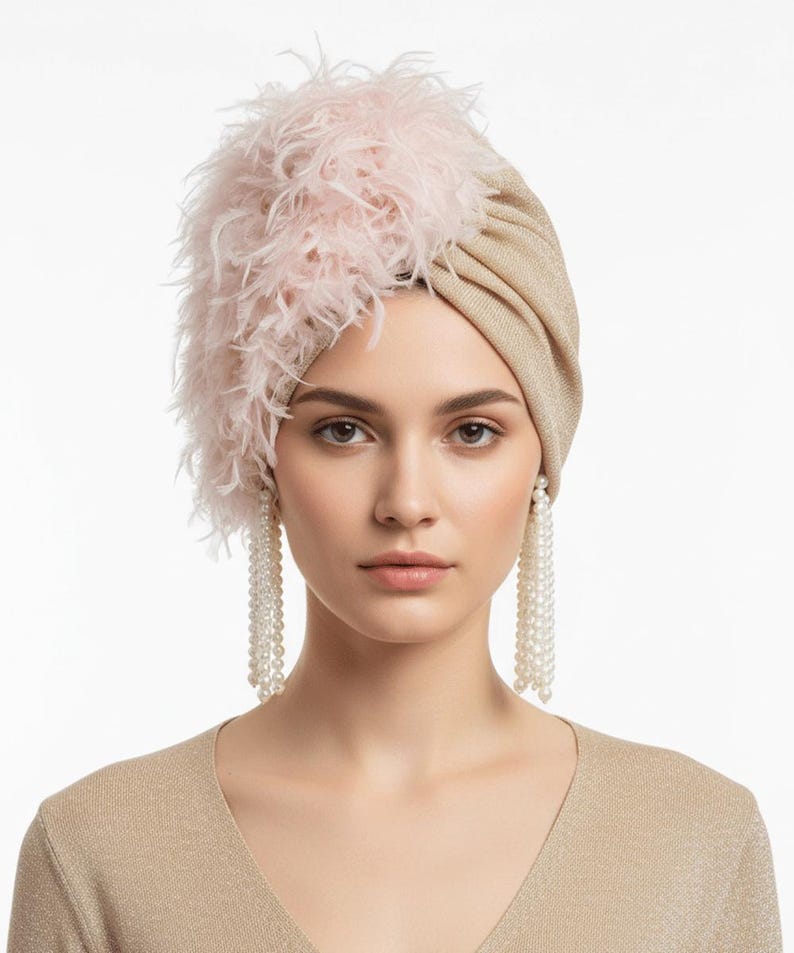 Puede incluir: Un turbante beige con un acento de plumas rosas. La mujer de la imagen lleva pendientes largos de perlas y una blusa beige. El turbante es un tocado elegante.