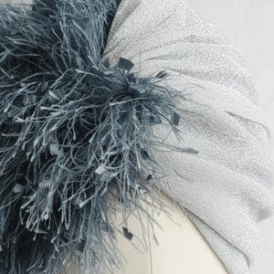 Puede incluir: Un turbante plateado con tela brillante, adornado con un grupo de plumas gris oscuro y peque&ntilde;os detalles cuadrados. Este tocado es un accesorio elegante, ideal para ocasiones especiales.