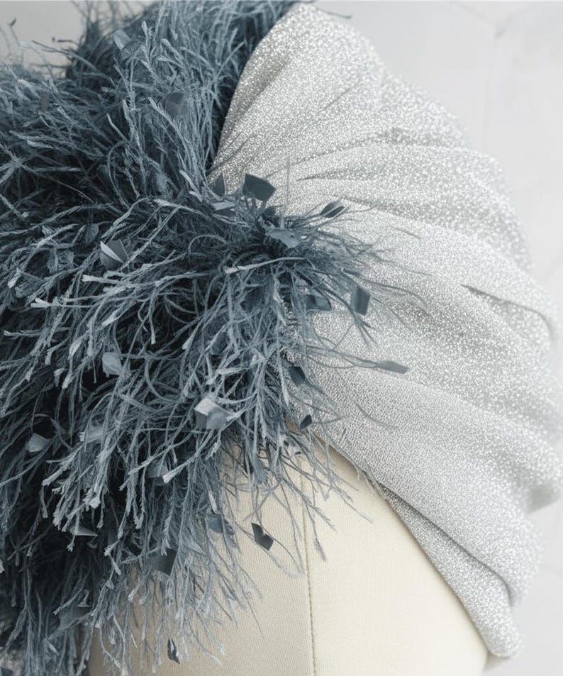 Puede incluir: Un turbante plateado con tela brillante, adornado con un grupo de plumas gris oscuro y peque&ntilde;os detalles cuadrados. Este tocado es un accesorio elegante, ideal para ocasiones especiales.