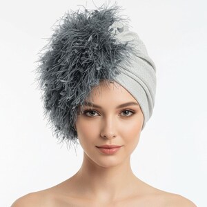 Puede incluir: Un turbante plateado con una apariencia texturizada y brillante. El turbante est&aacute; adornado con una gran disposici&oacute;n asim&eacute;trica de plumas grises en el lado izquierdo. El tocado lo lleva una persona de piel clara y ojos marrones.