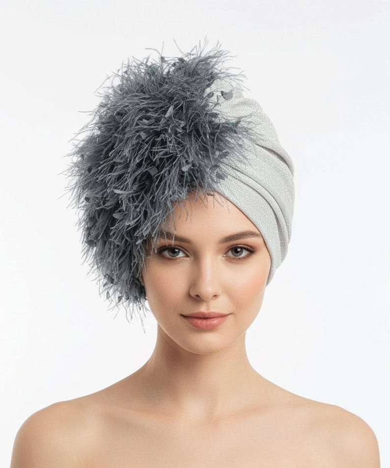 Puede incluir: Un turbante plateado con una apariencia texturizada y brillante. El turbante est&aacute; adornado con una gran disposici&oacute;n asim&eacute;trica de plumas grises en el lado izquierdo. El tocado lo lleva una persona de piel clara y ojos marrones.