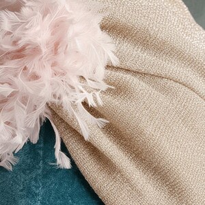 Puede incluir: Primer plano de un turbante dorado con tejido texturizado. El turbante est&aacute; adornado con un grupo de plumas rosas claras y blancas. El fondo es de tela color turquesa.