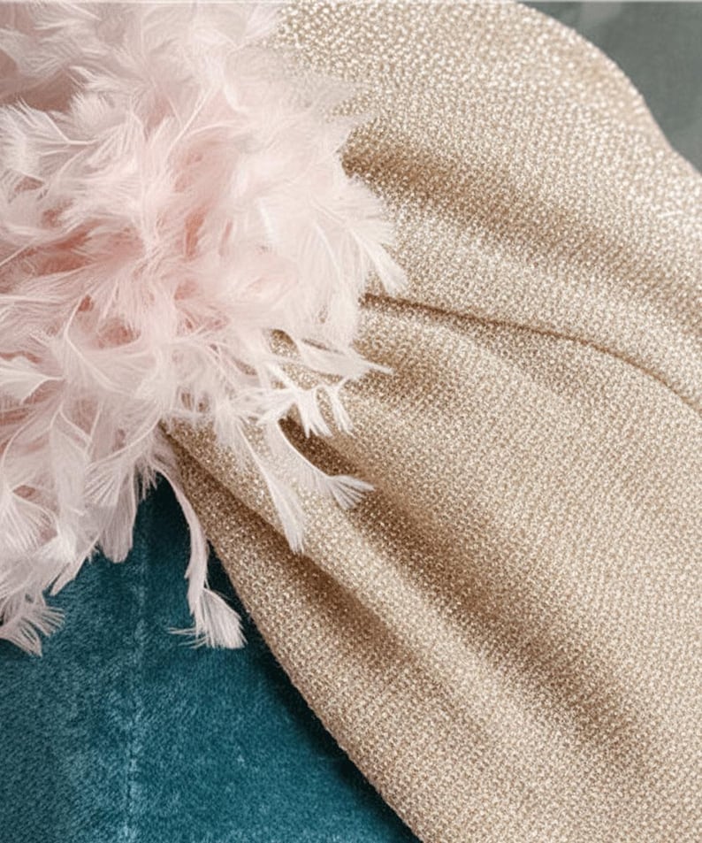 Puede incluir: Primer plano de un turbante dorado con tejido texturizado. El turbante est&aacute; adornado con un grupo de plumas rosas claras y blancas. El fondo es de tela color turquesa.