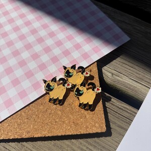 May include: Three enamel pins shaped like Siamese cats with tan bodies, dark brown faces, ears, and paws, and bright blue eyes. Each cat is depicted in a standing pose with a small white cloud and a red heart symbol positioned near the tail area.