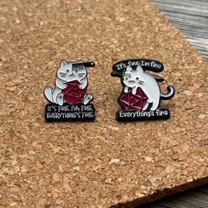 Pode incluir: Dois pins de esmalte com gatos brancos segurando dados vermelhos de vinte lados. O pin esquerdo mostra um gato com um cutelo na cabeça e o texto IT'S FINE. I'M FINE. EVERYTHING'S FINE. O direito mostra um gato com uma espada e o texto It's fine. I'm fine. Everything's fine.