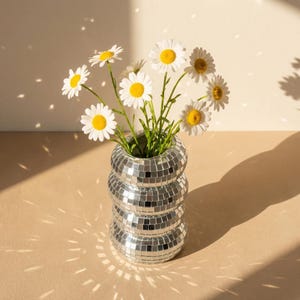 Può includere: Un vaso decorativo con un design a mosaico a specchio, che contiene un bouquet di margherite. Il vaso ha una forma unica e impilata. Le margherite hanno petali bianchi e centri gialli, con steli verdi.