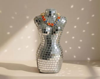 May include: A torso-shaped display mannequin covered in small, square, reflective mirror tiles. The mannequin is adorned with a necklace featuring alternating orange and blue circular stones set in a gold-toned metal chain.