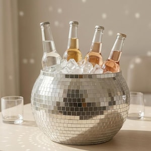 Op de afbeelding: Een zilveren ijsemmer in de vorm van een discobal, gevuld met ijs en vier glazen flessen. De flessen bevatten heldere, lichtgele en roze vloeistoffen. De emmer is rond en bedekt met kleine, reflecterende vierkantjes.