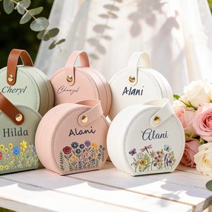Può includere: Una collezione di custodie rotonde color pastello personalizzate con manici. Ogni custodia è adornata con un nome e ricami floreali. Le custodie sono nei toni del rosa, verde e bianco, con manici marroni e bianchi. Un bouquet di rose rosa sullo sfondo.