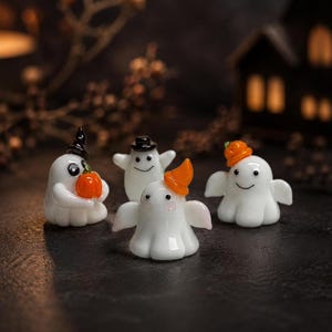 May include: Four small glass ghost figurines in white with black eyes and smiles. One ghost wears a black hat and holds an orange pumpkin, one wears a black top hat, one wears an orange hat, and one wears a small orange pumpkin on its head.