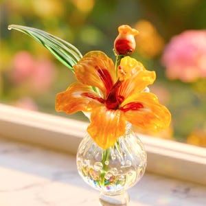 Puede incluir: Una escultura floral de vidrio hecha a mano con una flor de hibisco naranja con detalles rojos, una hoja verde y un capullo. La flor se exhibe en un jarrón de vidrio transparente con detalles de burbujas.