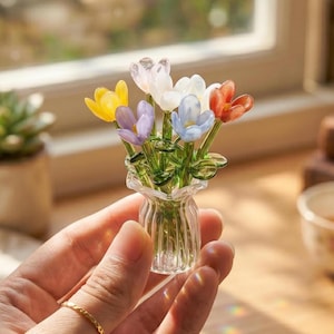 Op de afbeelding: Een kleine glazen vaas gevuld met kleurrijke glazen bloemen. De bloemen zijn in tinten geel, paars, wit, blauw en oranje. De vaas is helder en heeft een gerimpelde rand. De opstelling wordt in een hand gehouden.