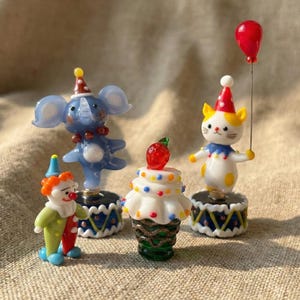 May include: A collection of whimsical circus-themed figurines. Includes a blue elephant, a clown, a cupcake with a strawberry, and a cat holding a red balloon. Each figurine is on a decorative base with a colorful design.