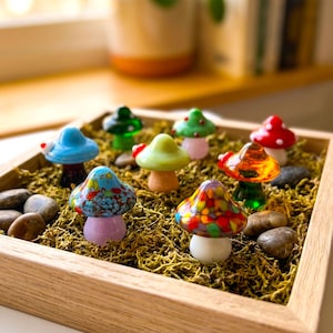 May include: A wooden tray filled with moss and small, colorful glass mushroom figurines. The mushrooms have various colors, including blue, green, red, and yellow, with speckled and solid designs. Small stones are scattered around the mushrooms.