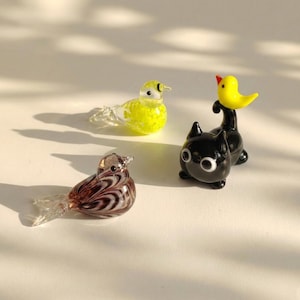 Pode incluir: Três figuras de vidro: um pássaro amarelo, um gato preto com cabeça de pato amarelo e um pássaro marrom e branco. As figuras têm asas e caudas de vidro transparente. Os objetos estão em um fundo branco com sombras.