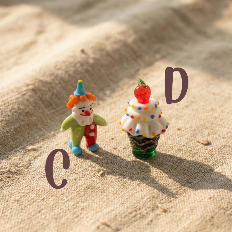 May include: Two miniature glass figurines: a clown and a cupcake. The clown is dressed in colorful attire with a blue hat. The cupcake has white frosting with colorful sprinkles and a red cherry on top. The figurines are on a tan surface.