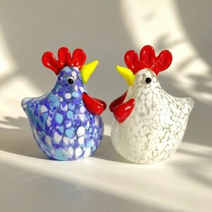May include: Two glass rooster figurines. One is blue with white and light blue spots, the other is white with gray spots. Both have red combs and wattles, and yellow beaks. These decorative figurines are perfect for a kitchen or farmhouse decor.