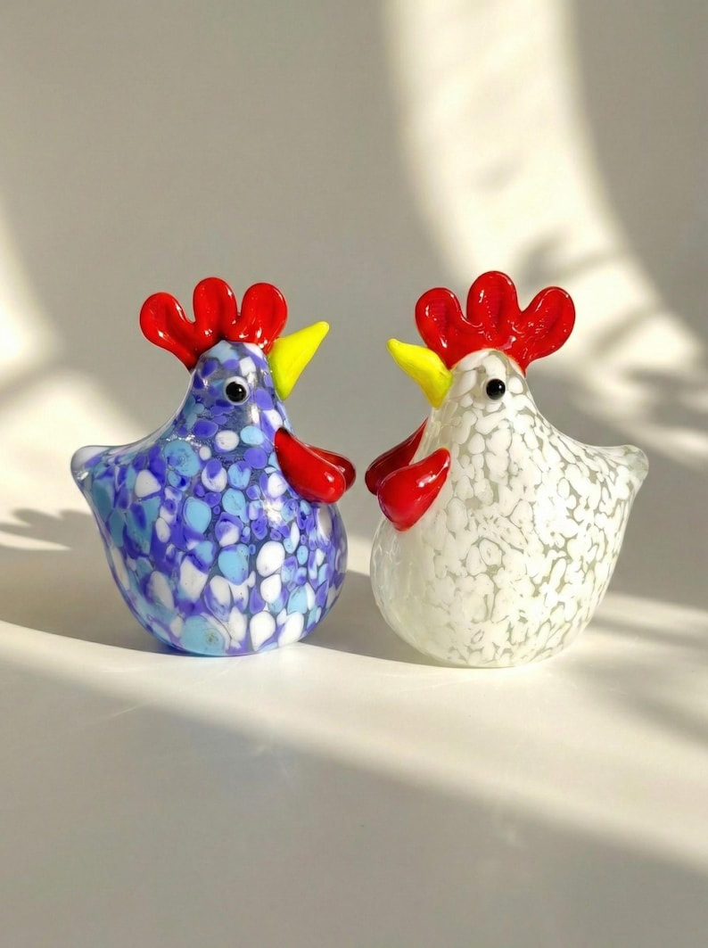 May include: Two glass rooster figurines. One is blue with white and light blue spots, the other is white with gray spots. Both have red combs and wattles, and yellow beaks. These decorative figurines are perfect for a kitchen or farmhouse decor.